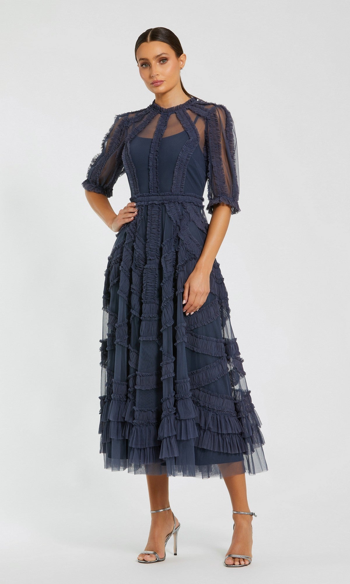 A woman in a Mac Duggal 8058 Ruffled Midi Wedding-Guest Dress in midnight with sheer sleeves and silver high-heeled sandals stands against a plain light background.